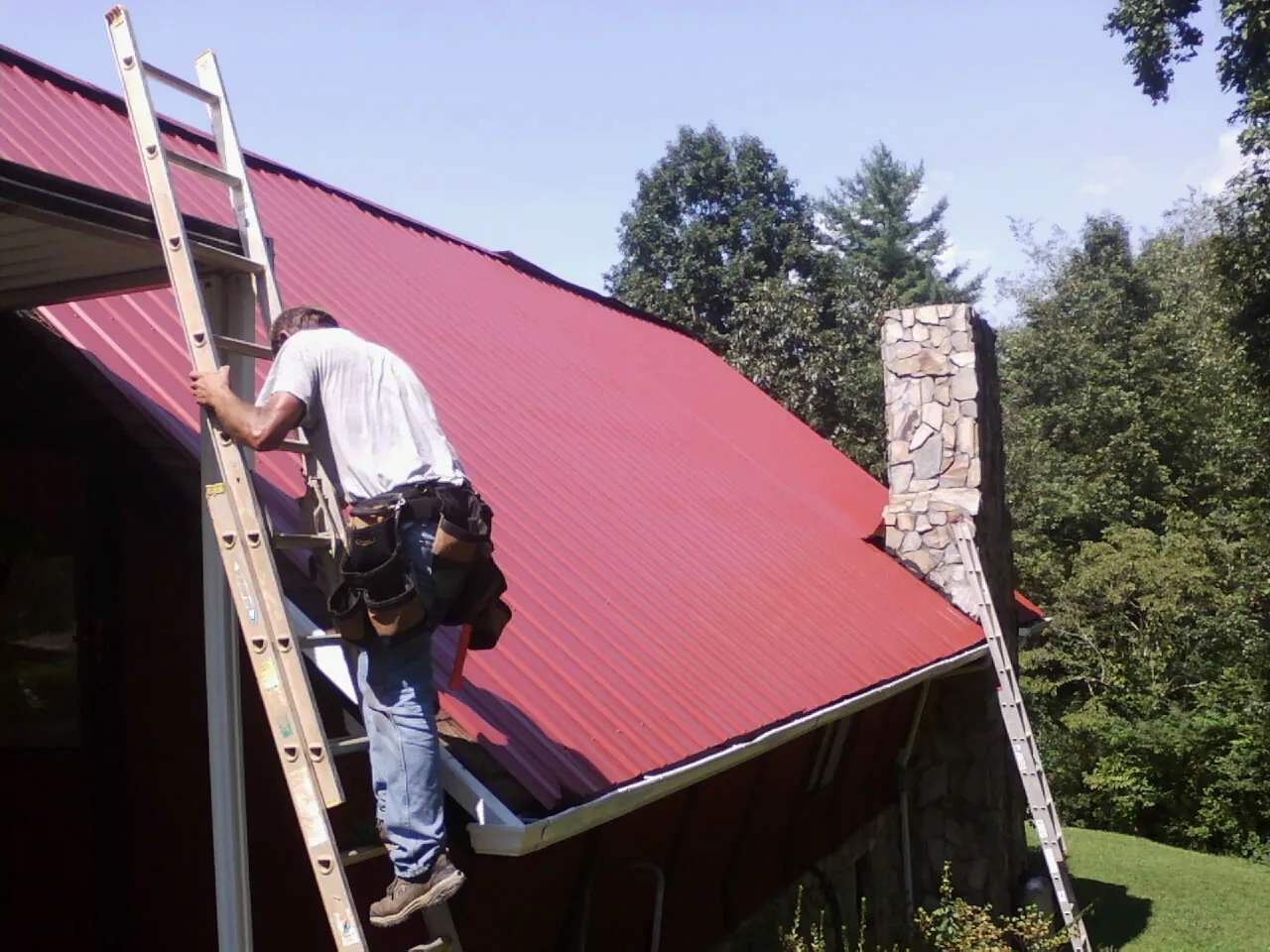 Roofer on a ladder beside a red metal roof and stone chimney — Soffit & Fascia Repair by Roofing Brothers in New Bedford